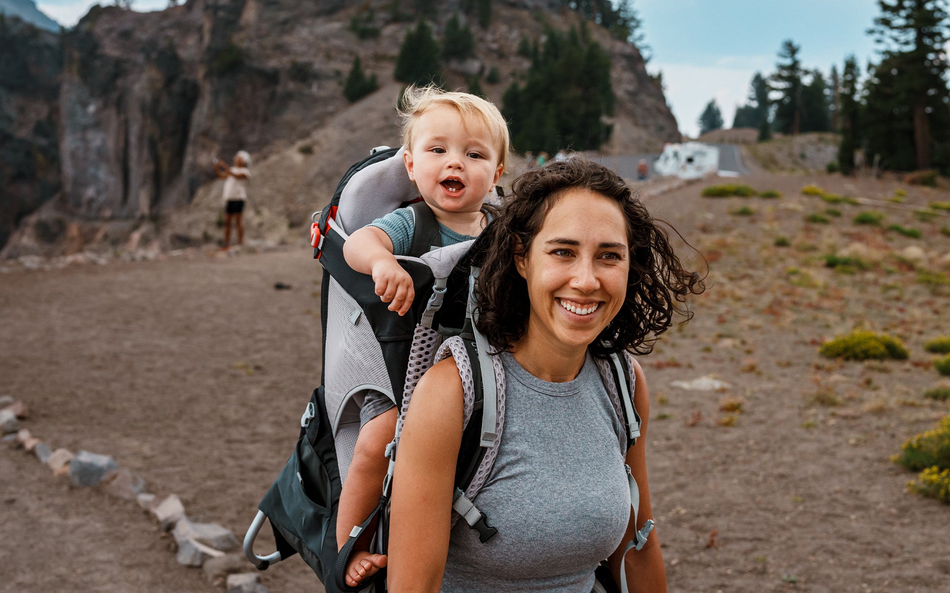 Woman smiling while carrying a baby on her back outdoors.